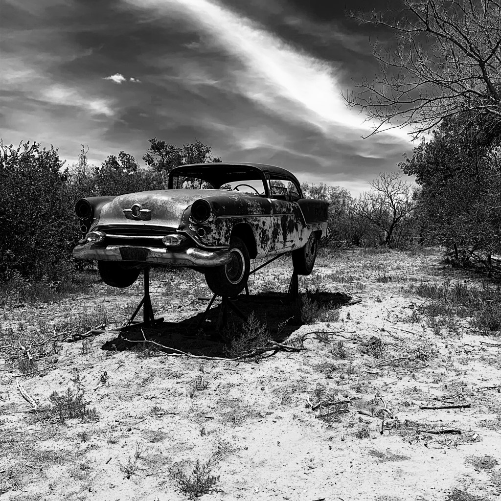 An old car sitting up on a rack in the middle of a mesquite pasture with clouds in the sky. Photo is in black and white. 
